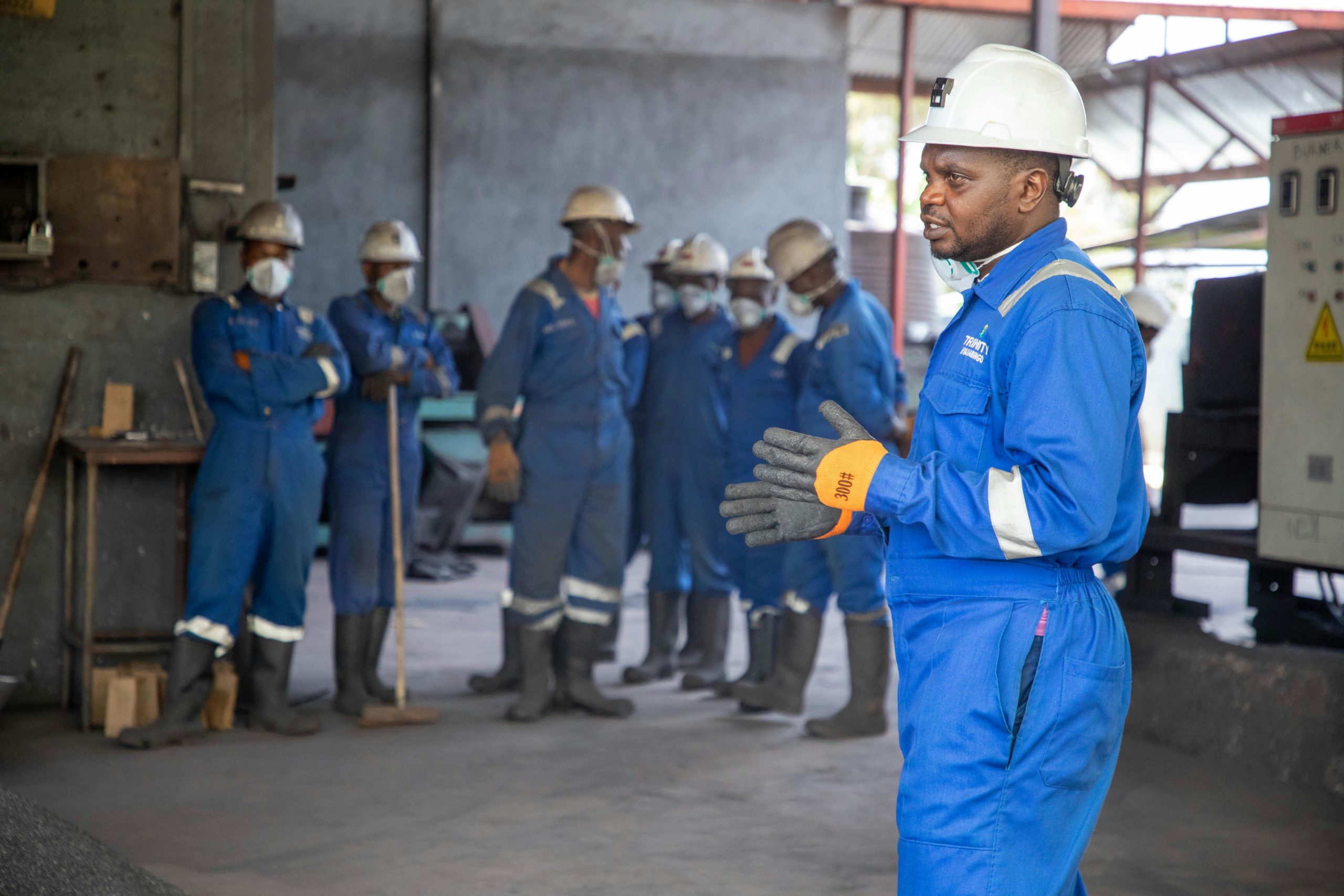 Homme en tenue de travail bleue et casque blanc, portant des gants, s’adressant à un groupe d’ouvriers équipés de casques et de protections, dans un atelier industriel.